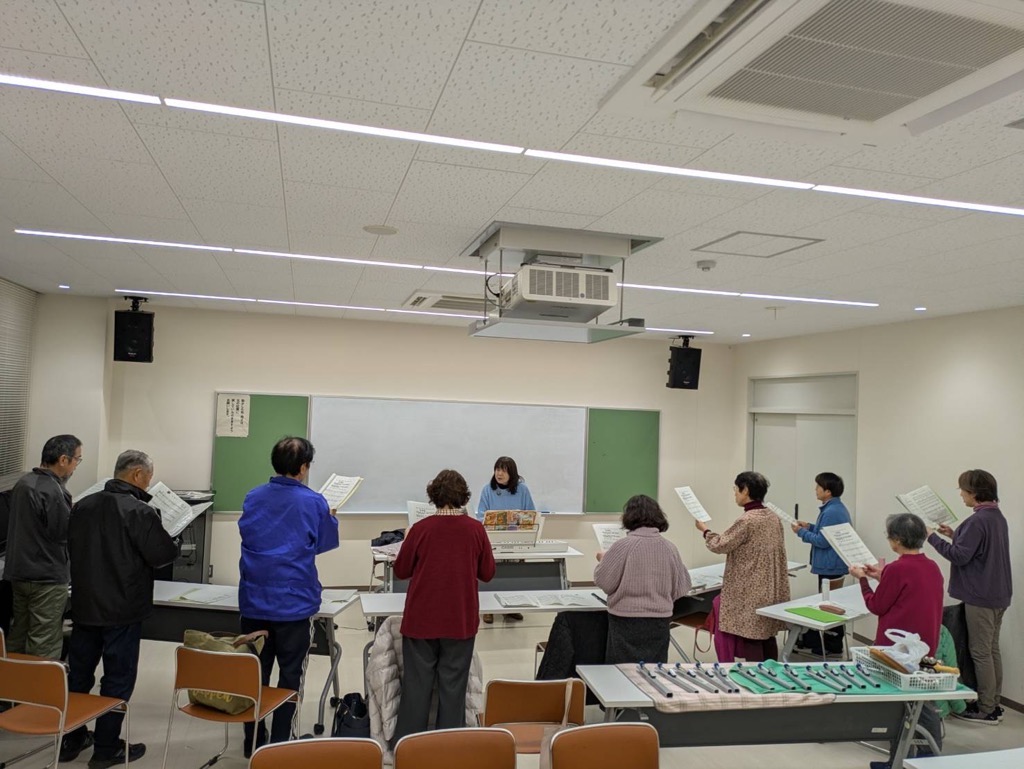 先生を囲んで生徒が楽譜を持ち歌の練習をしている写真