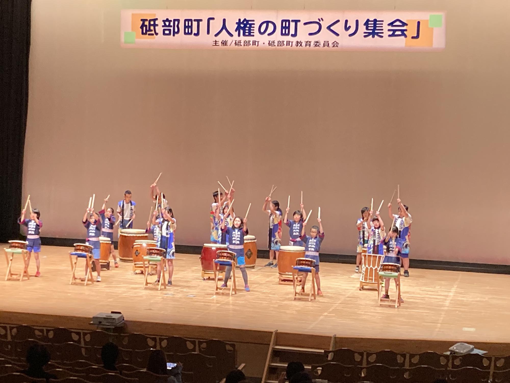 ホールの壇上で小学生が出演者が太鼓の演奏をしている写真