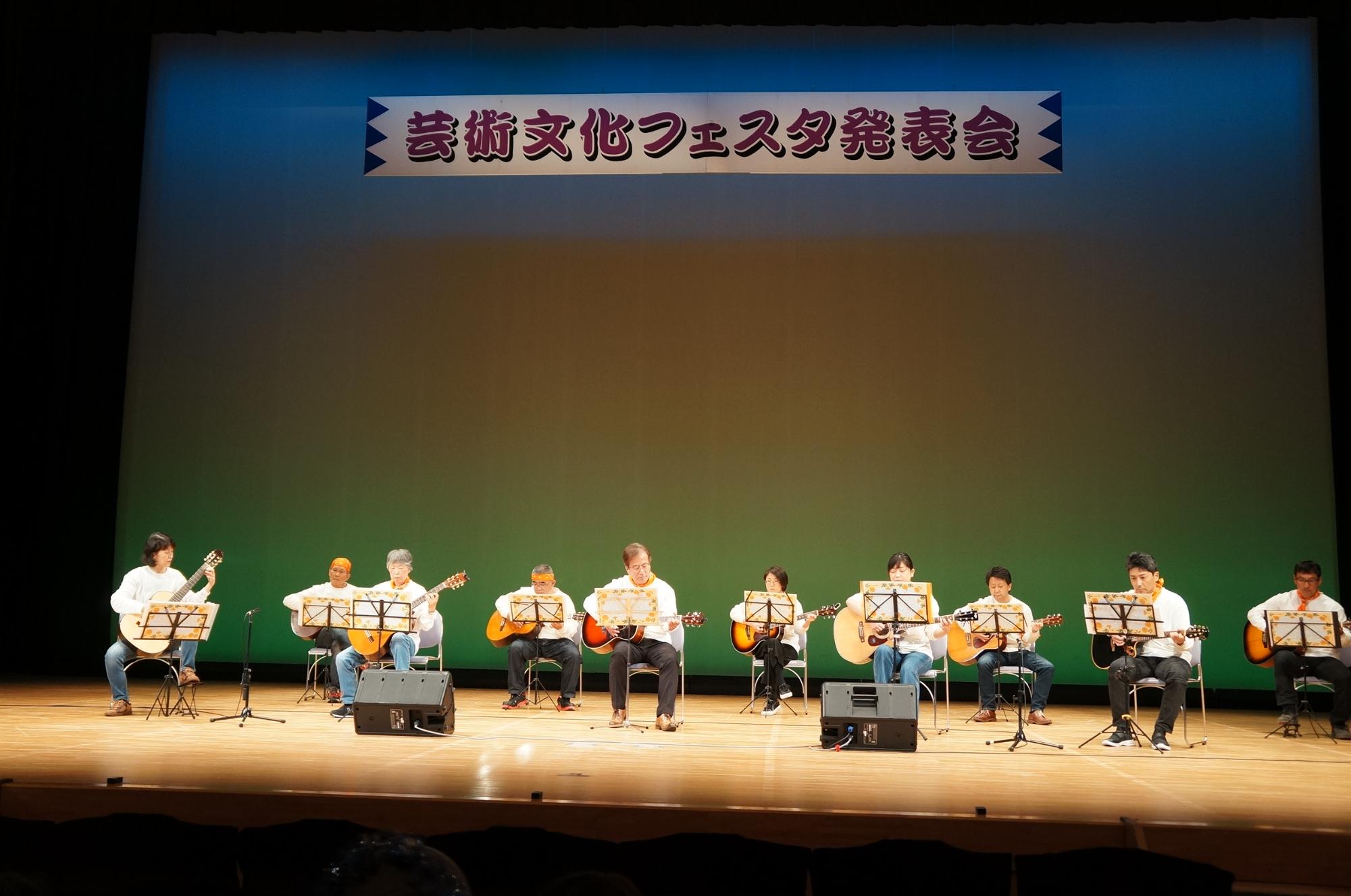 ステージでたくさんの人が椅子に座ってギターを演奏している写真